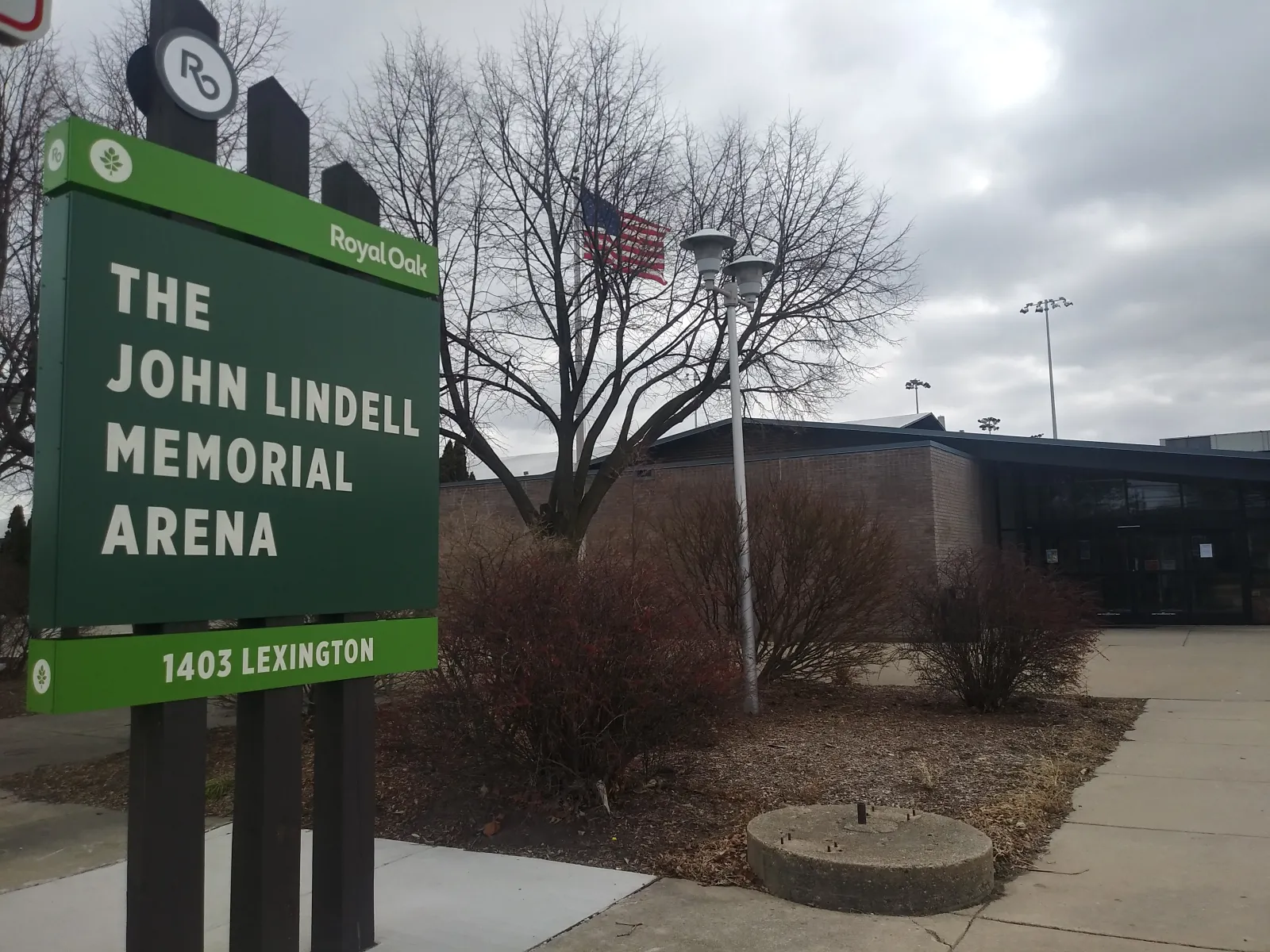 The green City of Royal Oak sign reading The John Lindell Memorial Arena, 1403 Lexington, in front of the low brick entrance building on an overcast day. — John Lindell Ice Arena