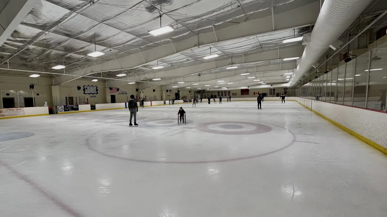 Full ice sheet inside the Line Creek arena during public skating, with curling circles painted on the ice, an American flag and Canadian flag on the wall, and scattered skaters — Line Creek Community Center and Ice Arena