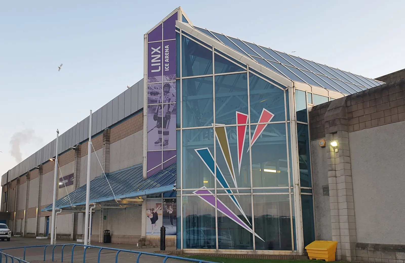 The glazed entrance tower with the purple LINX ICE ARENA banner, and the long brick-and-metal side of the arena. — Linx Ice Arena
