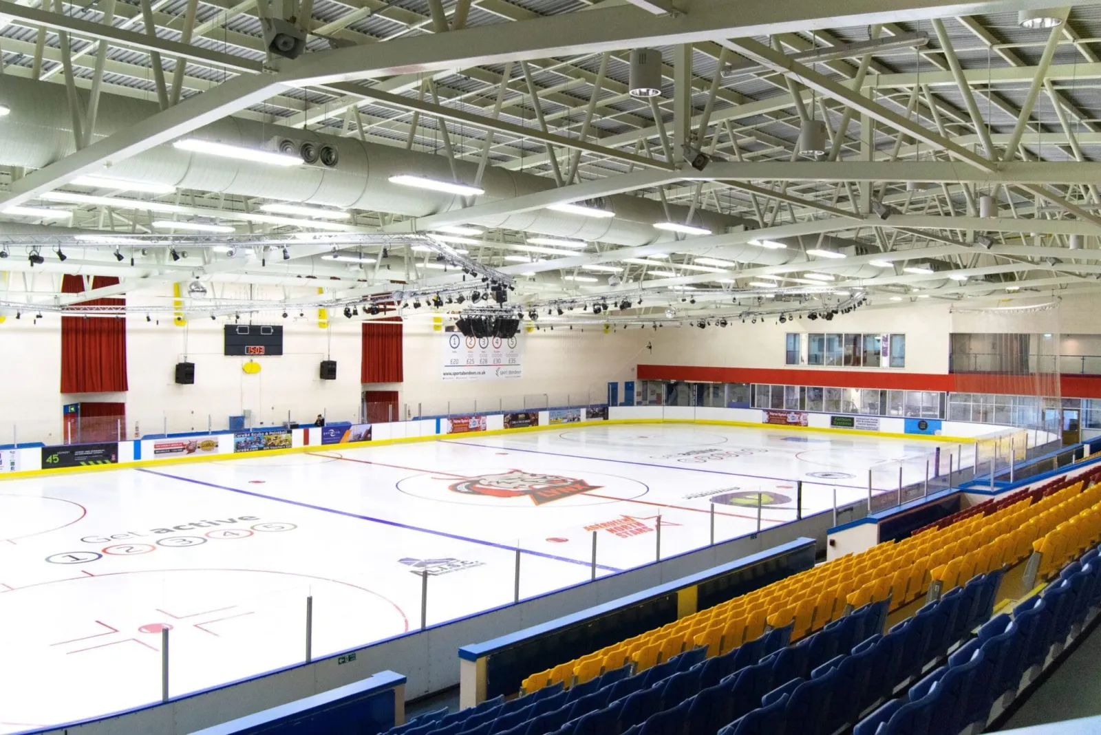 The empty ice from the seats, with the blue and yellow seating, scoreboard and steel roof trusses above. — Linx Ice Arena