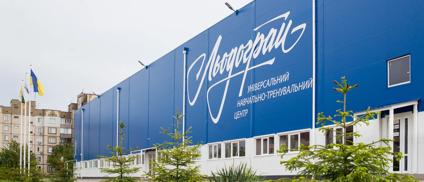 The long blue facade of the Liodohrai winter sports centre in Bohuslav with its large script logo, a Ukrainian flag at the corner and young pines along the front — Liodohrai Ice Arena Bohuslav