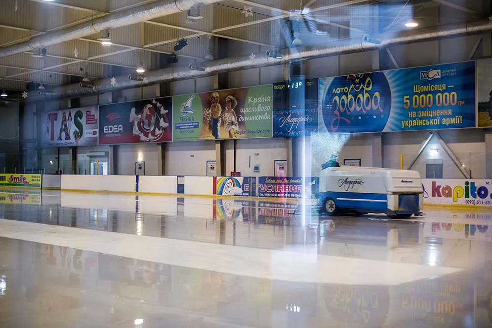 The empty ice sheet at Liodohrai with an ice resurfacer parked at the far end, white boards with sponsor banners along the sides and bright overhead lights — Liodohrai Ice Arena Bohuslav