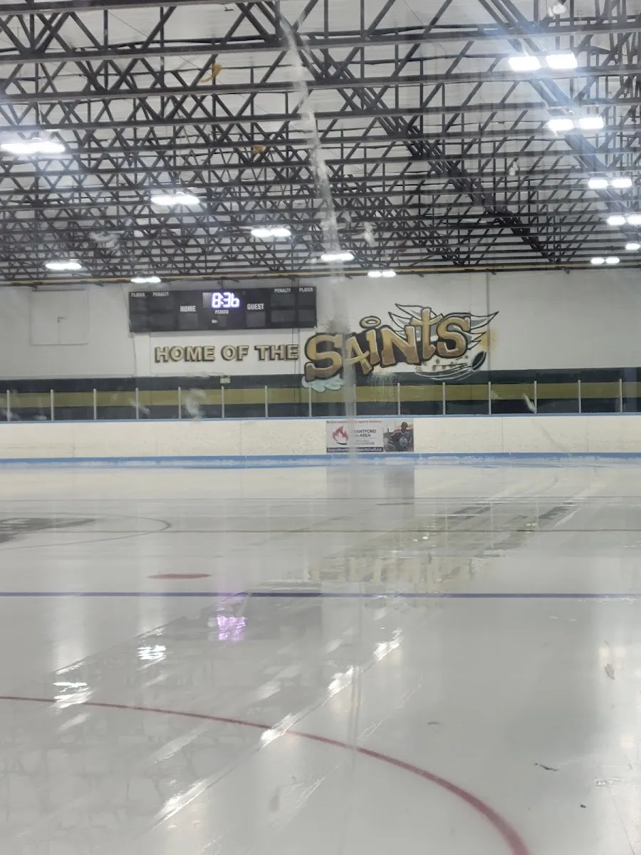 The empty ice at Lions Park Arena under a steel-truss roof, with a scoreboard and a Home of the Saints sign on the far wall. — Lions Park Arena