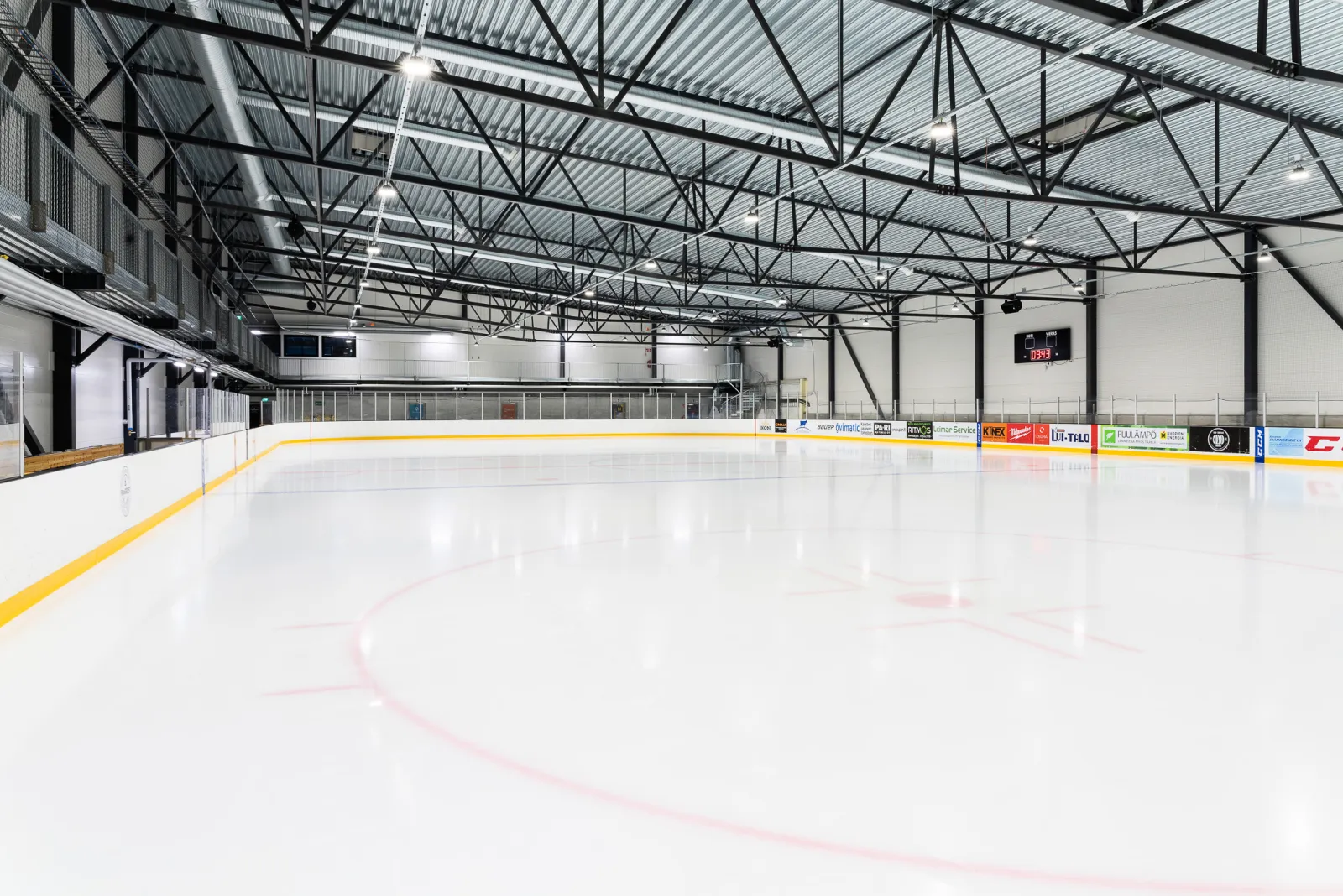 Den tomma isen i Lippumäki ishall under de svarta stålfackverken, sedd från hörnet med resultattavlan och sargen längs bortre långsidan – Lippumäen jäähalli (Tuplajäät)