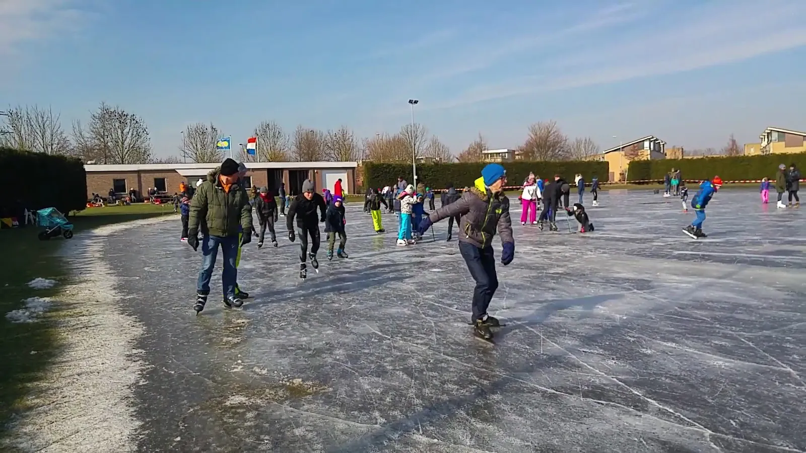 Schaatsers verspreid over het natuurijs van de Lisser baan op een zonnige winterdag, met het clubgebouw en hagen langs de verre rand. – Lissesche IJsclub