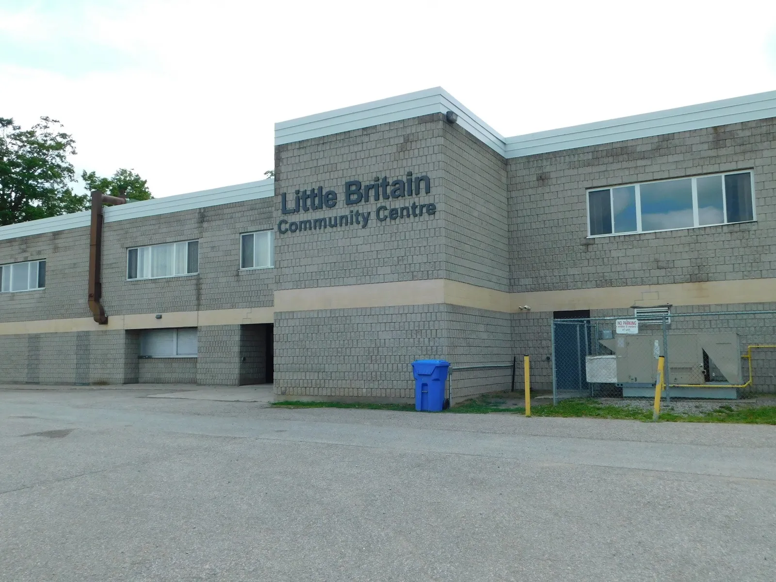 El Little Britain Community Centre, un edificio de dos plantas de bloques de hormigón gris con el nombre del centro en letras oscuras en la pared superior, visto desde el aparcamiento. — Little Britain Community Centre