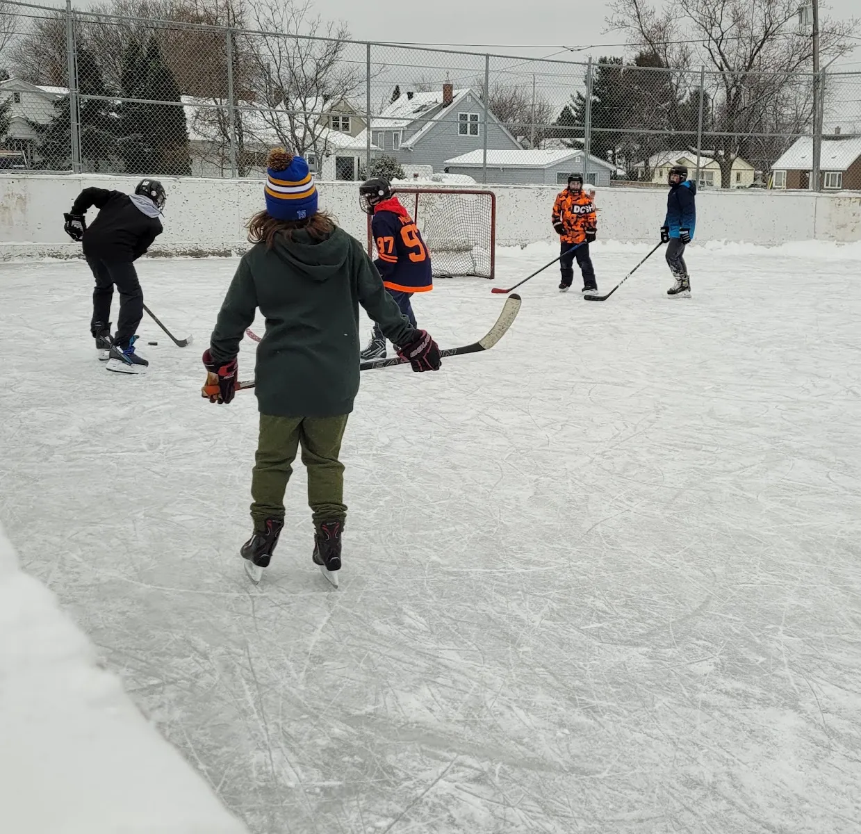 A pickup hockey game on the boarded outdoor rink, seen from the boards. — Lively Playground Outdoor Rink