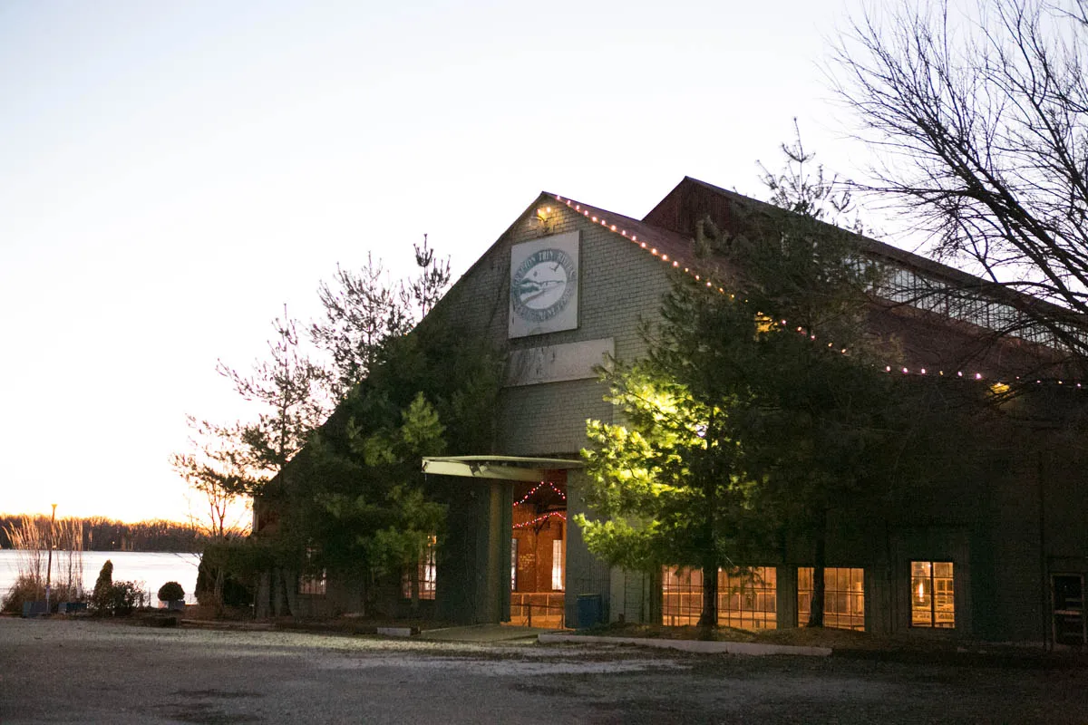 The Boatworks building at dusk, its gable roof outlined in lights, with the river behind the trees on the left. — The Loading Dock Ice Rink
