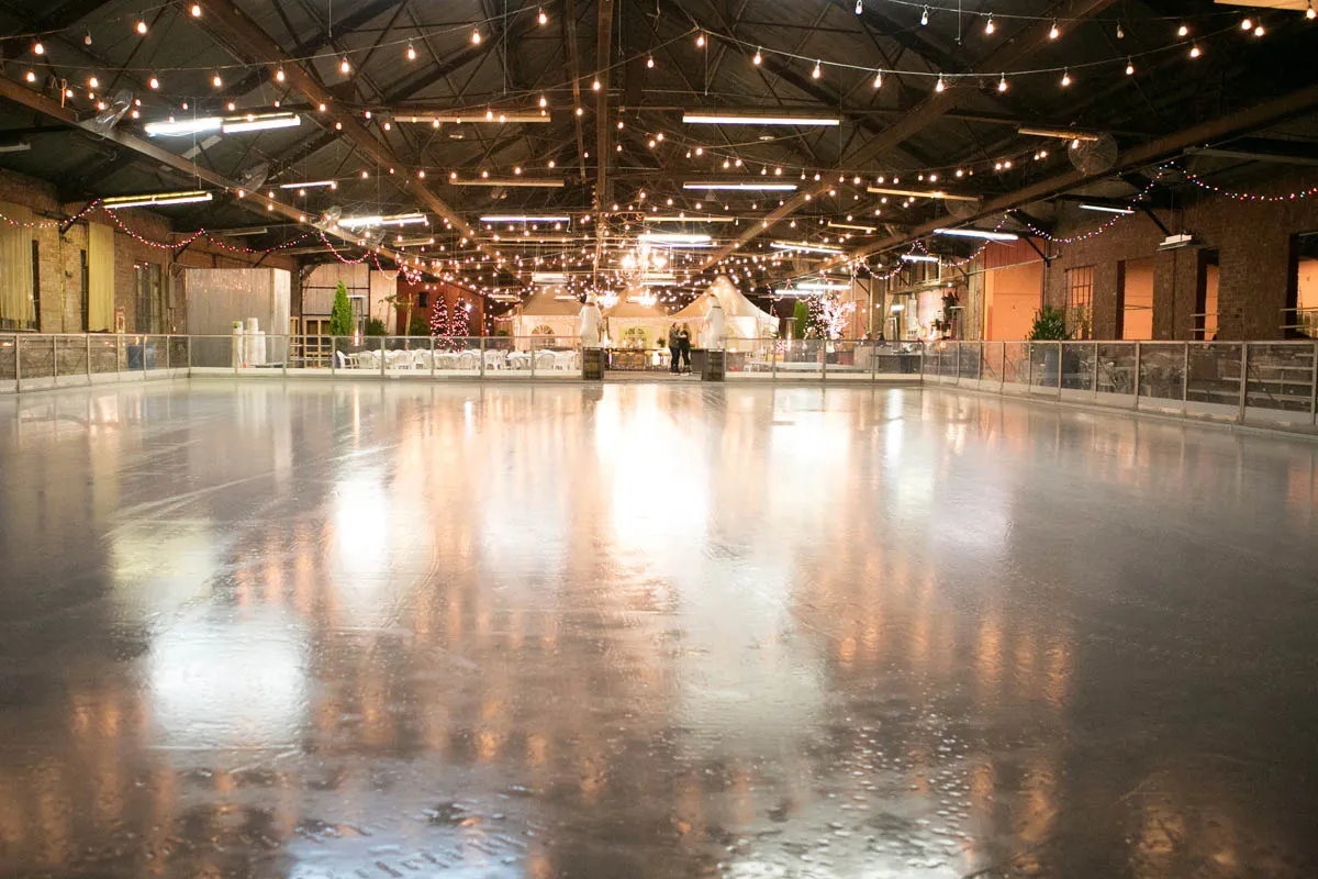 The empty rink inside the Boatworks under strings of lights hung from the steel roof trusses, with brick walls behind the boards. — The Loading Dock Ice Rink