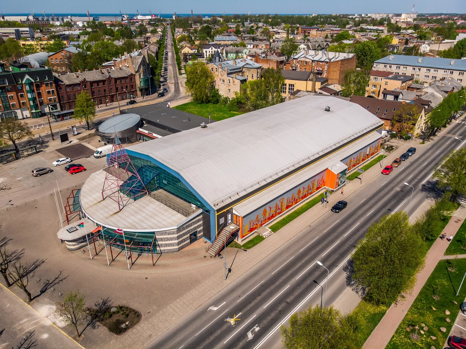 The ice hall seen from above and across the street, a long low building with a curved grey roof and a glazed corner entrance. — LOC Ledus halle