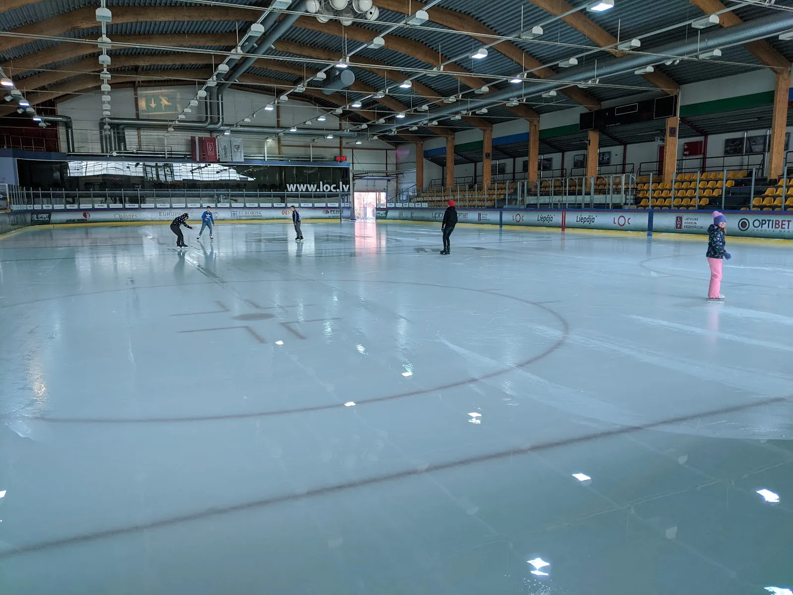 The ice sheet of the hall with a few skaters, the stands and the timber roof structure behind. — LOC Ledus halle