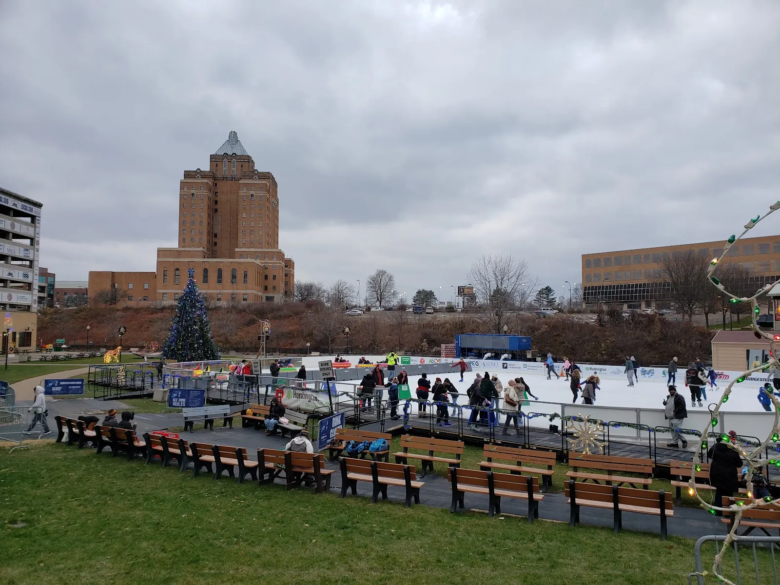 The seasonal rink at the far side of Lock 3 Park beneath the old Akron city skyline, with a lit Christmas tree, benches and skaters along the ice. — Lock 3 Ice Rinks