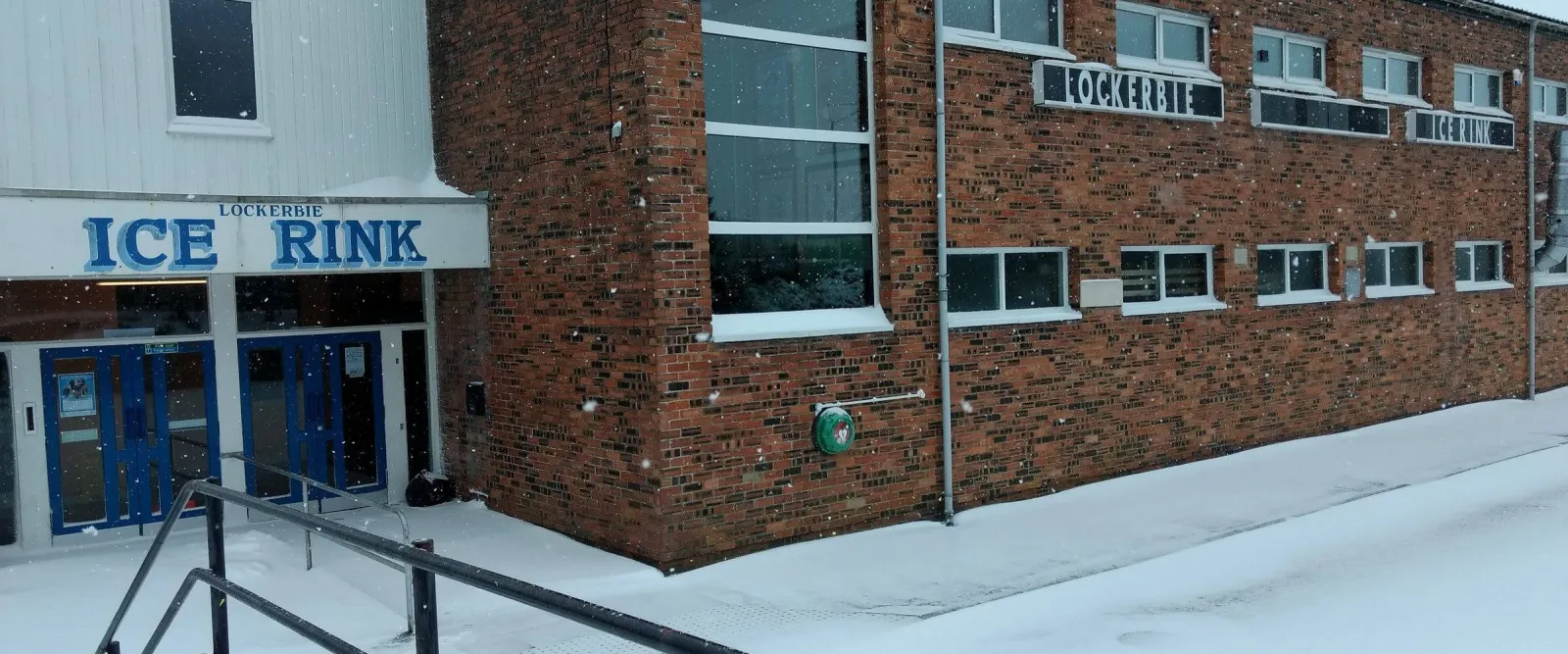 The red-brick front of the rink with its blue ICE RINK sign over the entrance, in snow. — Lockerbie Ice Rink