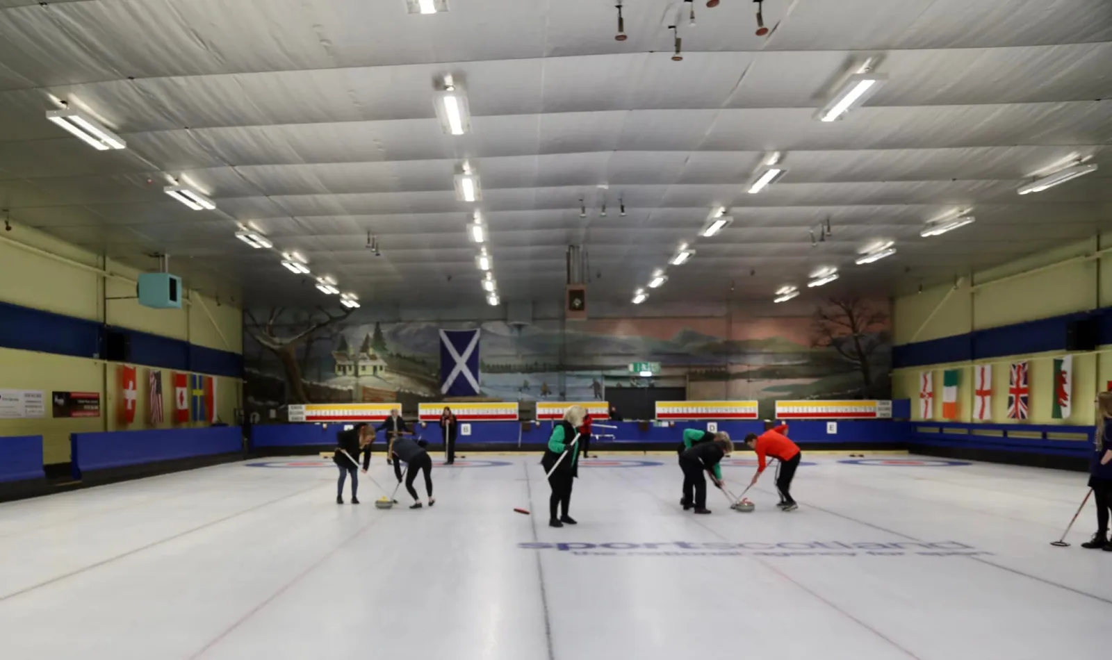 The long sheet of ice under the lit ceiling with a few curlers at the far end and flags along the back wall. — Lockerbie Ice Rink