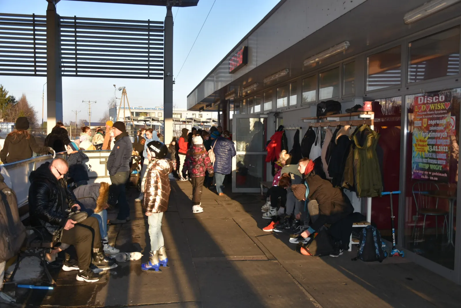The skate-hire kiosk with coat racks and benches beside the Żyrardów rink on a winter afternoon, with skaters queuing and lacing up under the roof overhang and a housing estate behind. — Lodowisko AQUA Żyrardów