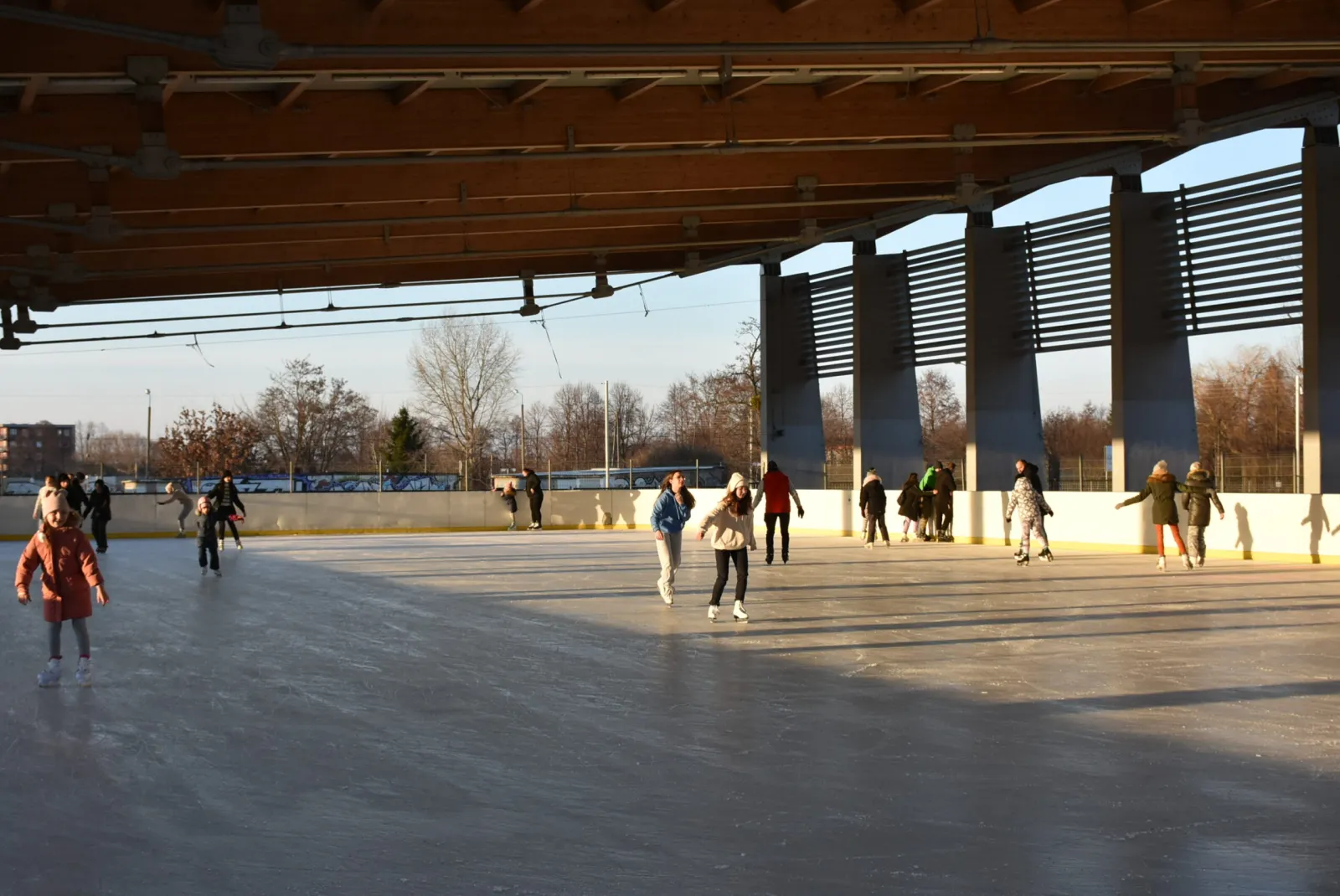 Skaters spread across the boarded ice of the Żyrardów rink under its timber-beamed open-sided roof, with concrete pillars, bare trees and apartment blocks beyond. — Lodowisko AQUA Żyrardów
