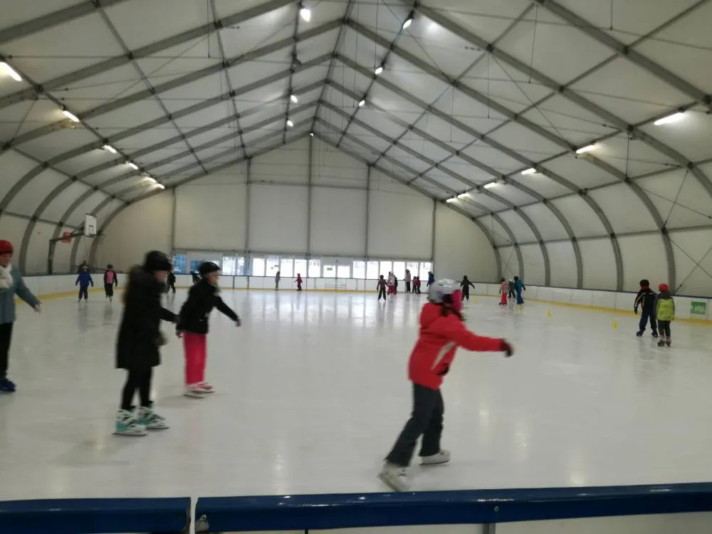 Skaters on the boarded ice inside the long white steel-framed tent hall of the Orlik seasonal rink, with overhead floodlights, advertising boards along the far side and a few families near the boards. — Sezonowe Lodowisko Kompleks Sportowy Orlik Bielsko-Biała