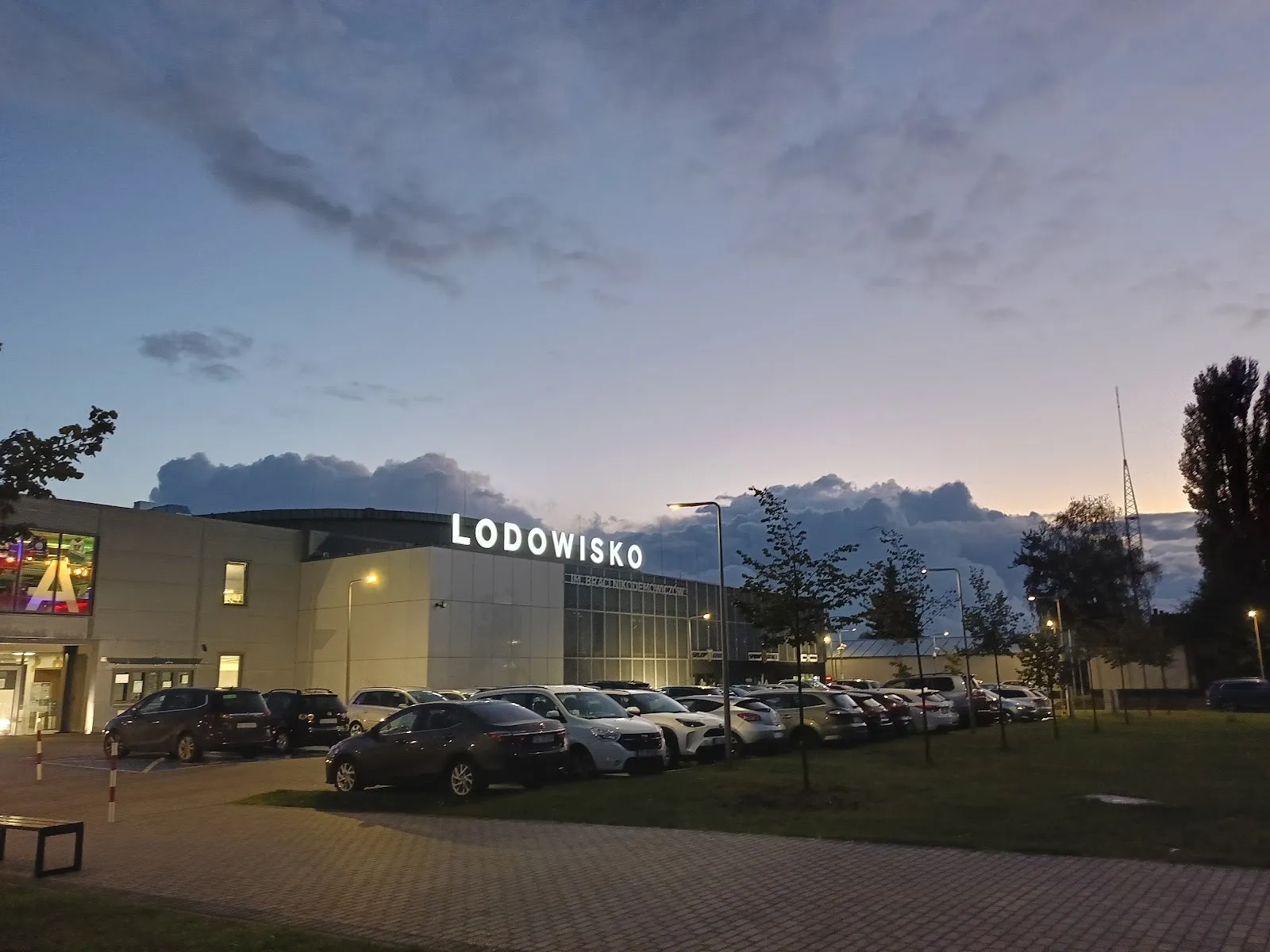 The Lodowisko ice rink building in Bytom at dusk, with its lit sign and the cars parked in front. — Lodowisko im. Braci Nikodemowiczów Bytom