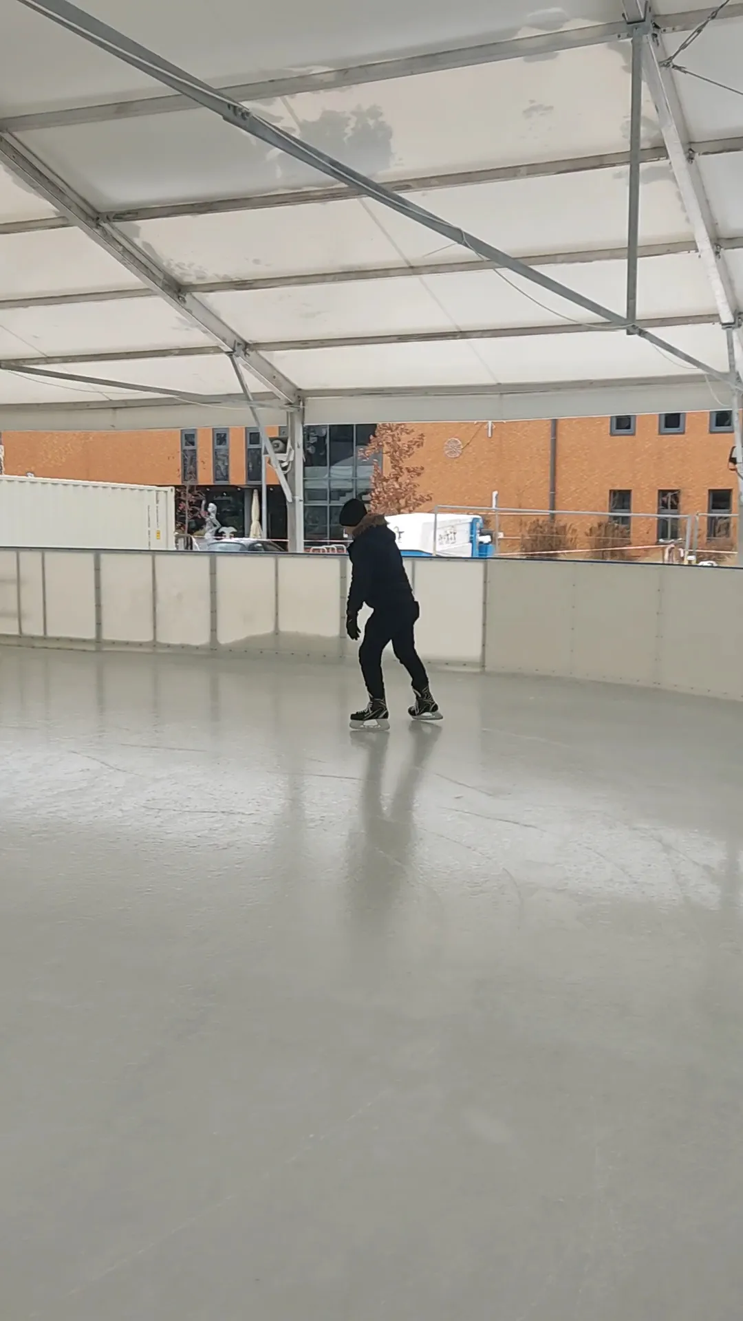 Samotny łyżwiarz na tafli zadaszonego lodowiska pod białym dachem namiotu, z budynkiem domu kultury widocznym za bandą. — Lodowisko Ursynów (Dzielnicowy Ośrodek Kultury, Kajakowa)