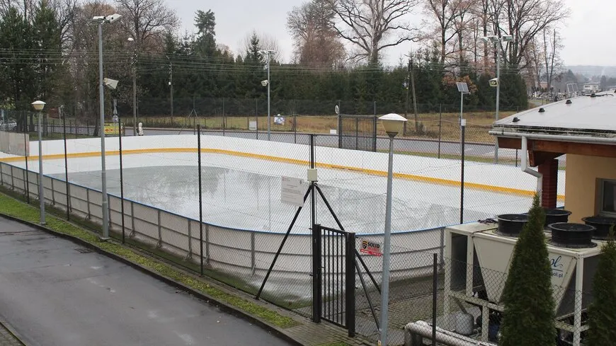 The empty ice sheet of the Jedlicze municipal rink behind a mesh fence, with floodlight poles, the refrigeration plant and trees beyond. — Lodowisko GOSiR Jedlicze
