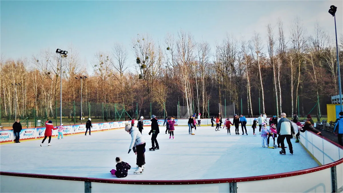 The open-air ice rink at ORS Żabka in Łaziska Górne on a winter afternoon, with skaters of all ages spread across the ice under floodlights and bare trees behind. — Lodowisko Łaziska Górne