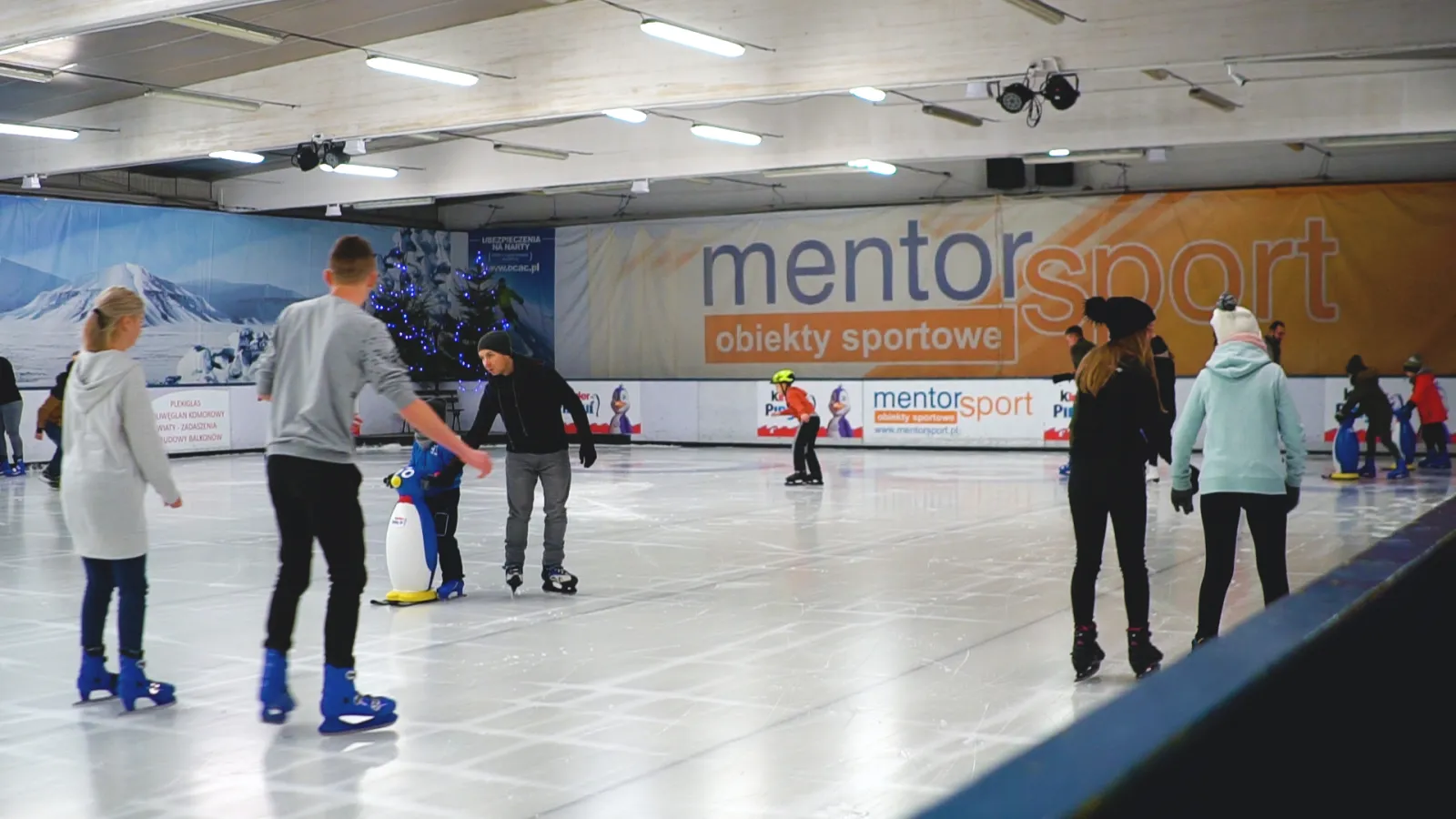 Łyżwiarze na krytym lodzie lodowiska MentorSport w Toruniu podczas ślizgawki, z banerami operatora wzdłuż band — Lodowisko MentorSport Toruń