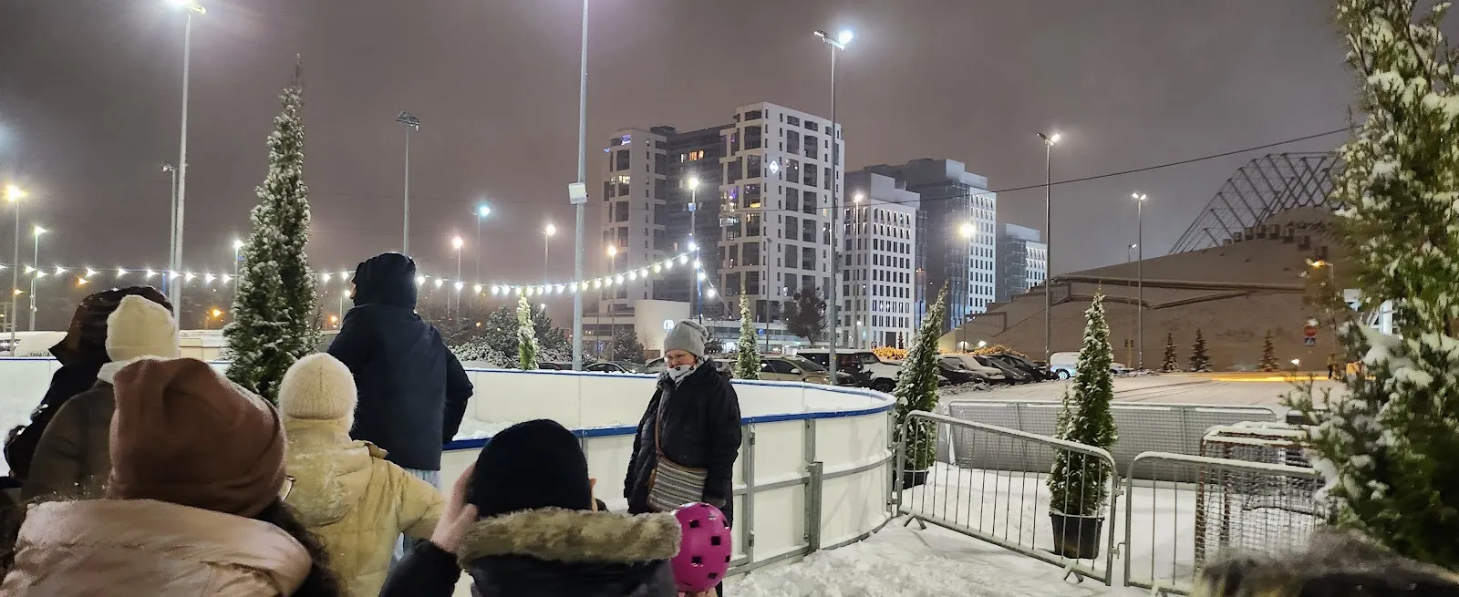 The rink's boarded edge seen from outside on a winter evening, with spectators at the entrance, fir trees and the Gdynia Arena roof behind. — Gdyńskie Lodowisko (Gdynia Arena)