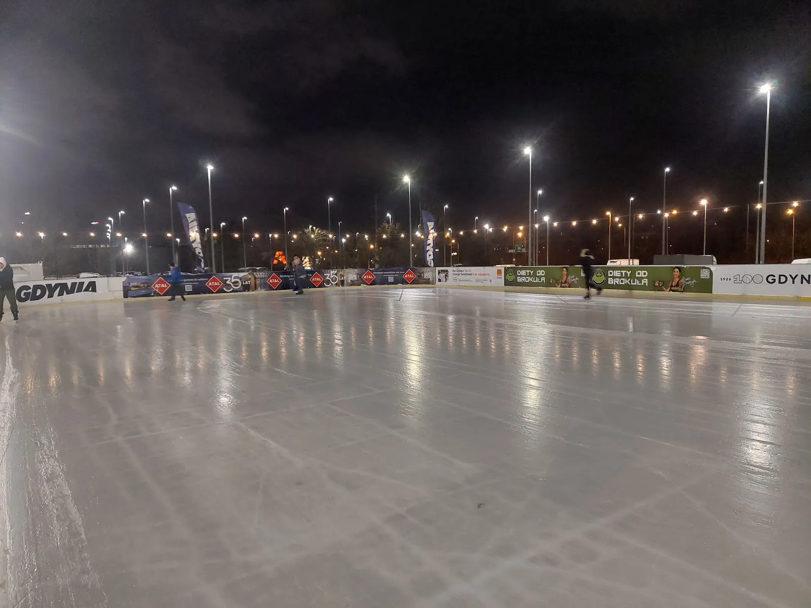 The empty ice sheet of the Gdynia city rink after dark, with the GDYNIA boards and the floodlights of the car park around it. — Gdyńskie Lodowisko (Gdynia Arena)