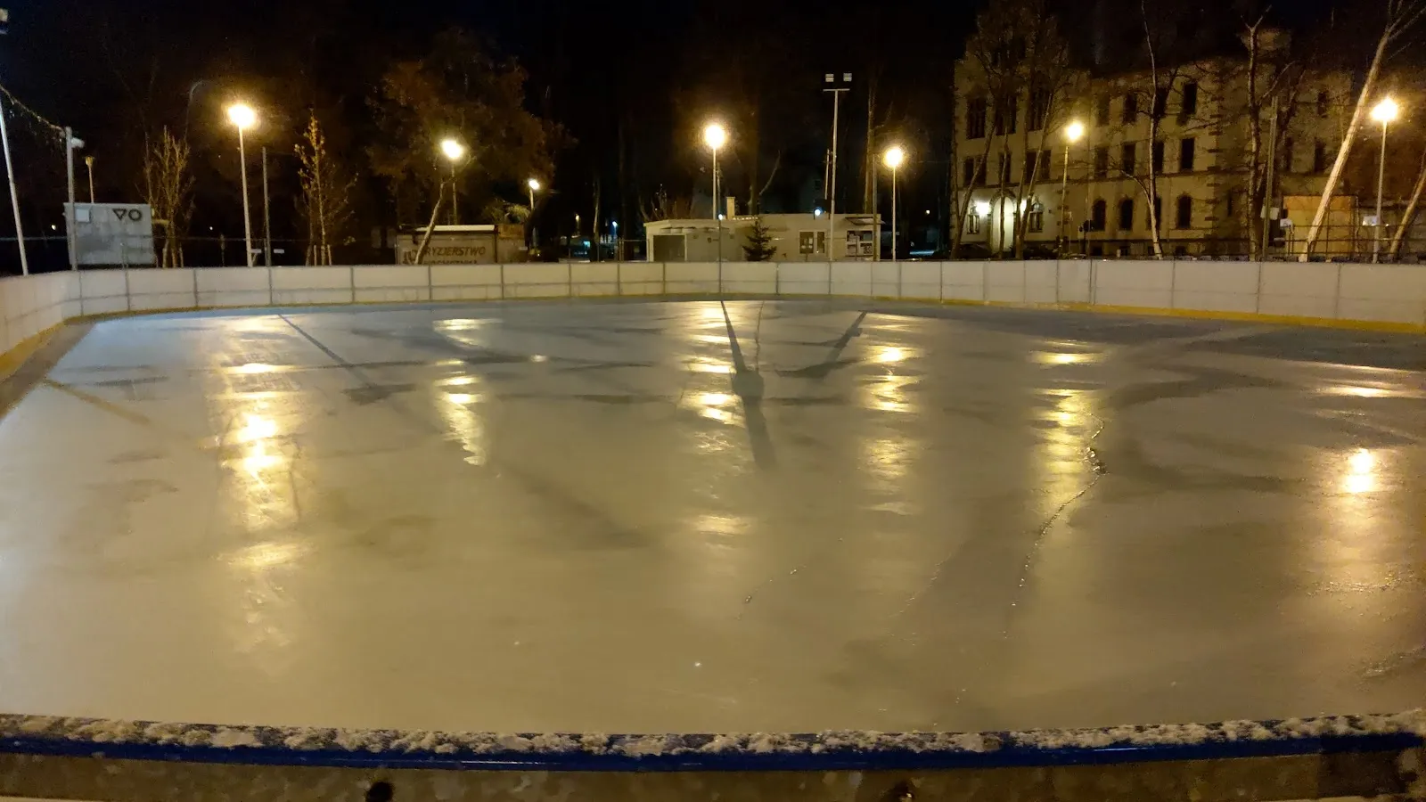 The empty ice of the Lubliniec city rink at night with its boards and lamps — Lodowisko Miejskie Lubliniec