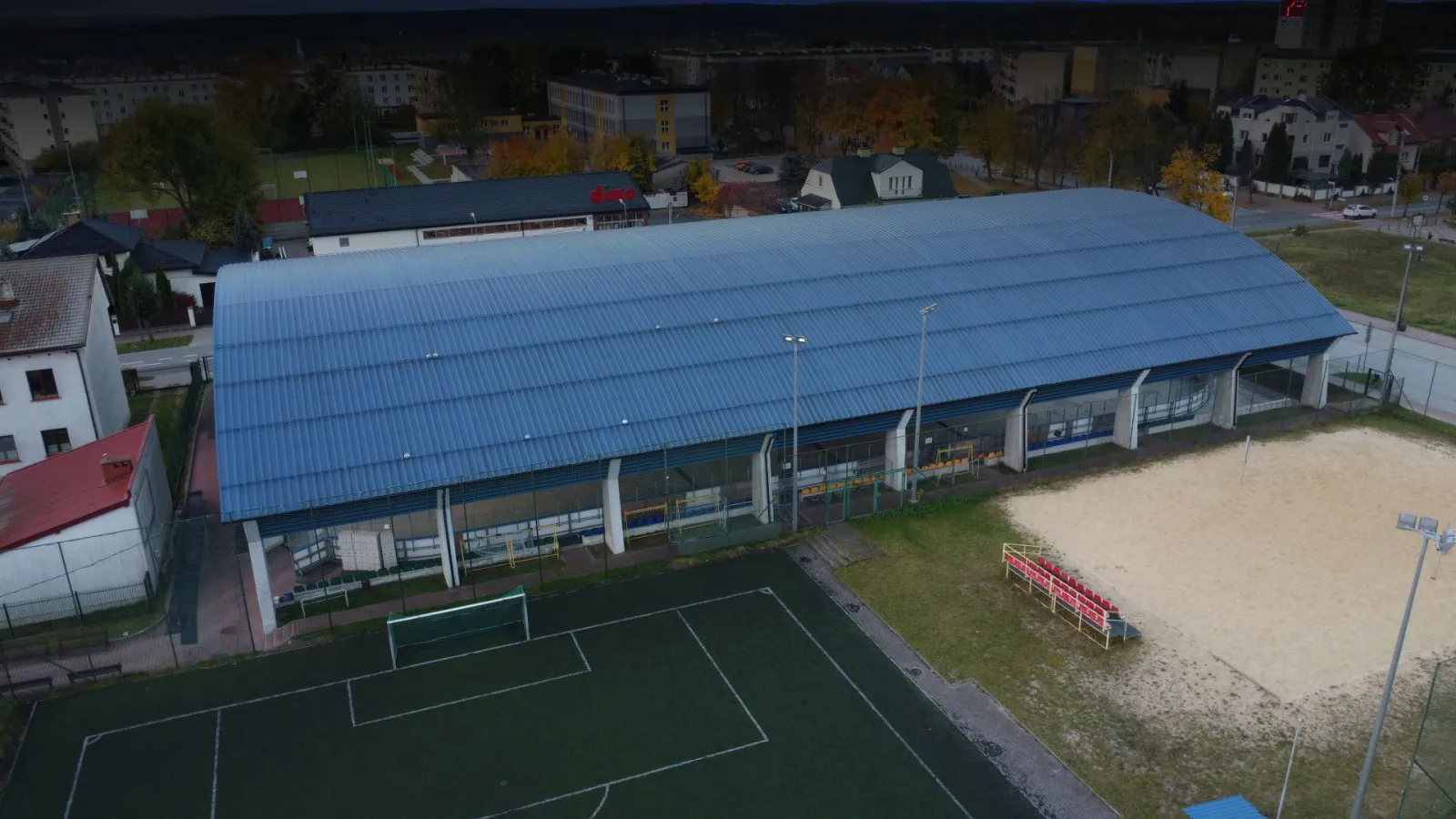 Aerial view of the Skarżysko-Kamienna rink hall: a long blue-roofed building with glazed side walls beside a sand volleyball court and a football pitch, with housing and trees beyond. — Lodowisko Miejskie Skarżysko-Kamienna