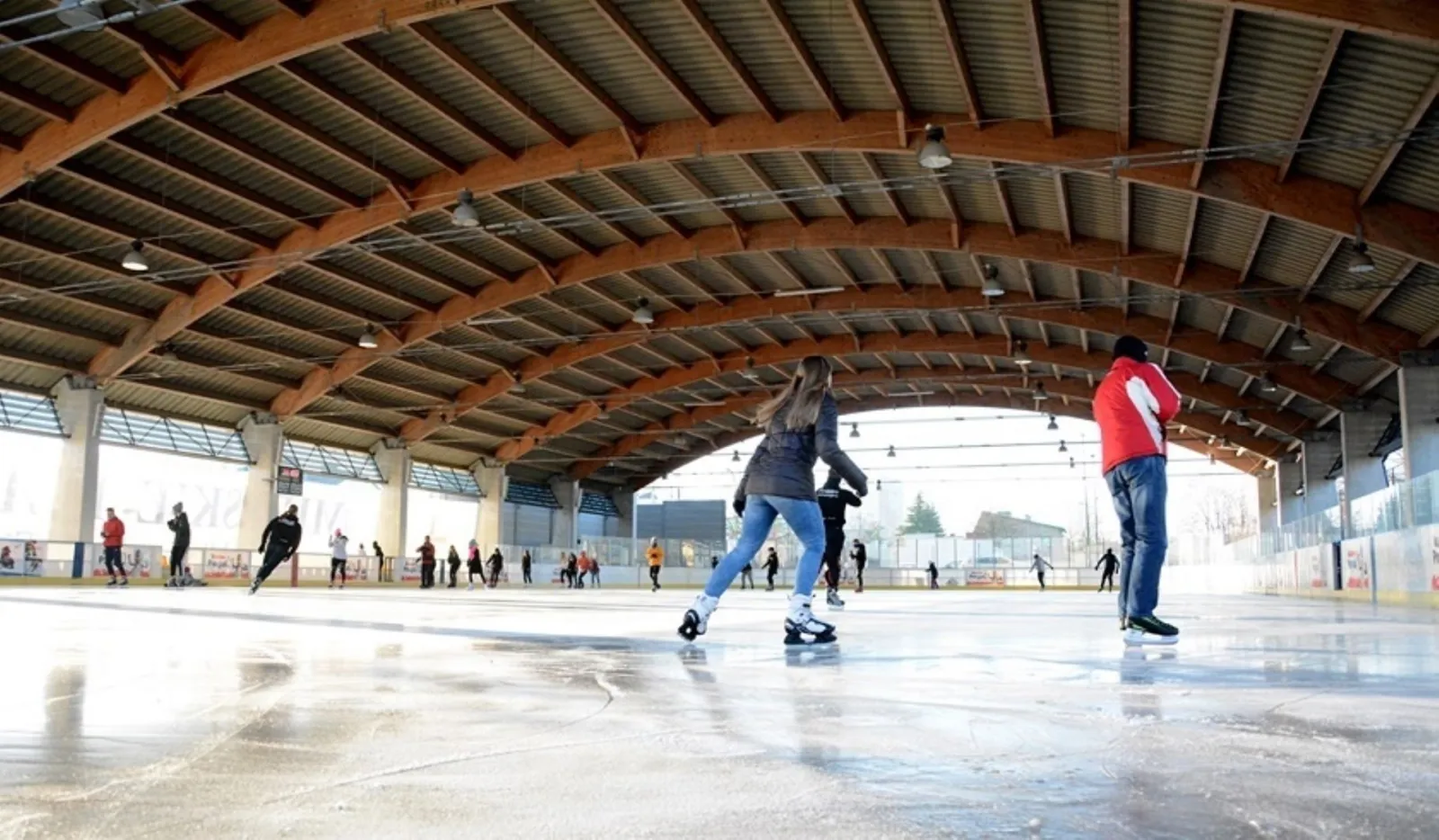 Skaters on the ice under the curved timber roof of the open-sided Skarżysko-Kamienna rink, with the boards and a scoreboard along the far side in winter daylight. — Lodowisko Miejskie Skarżysko-Kamienna