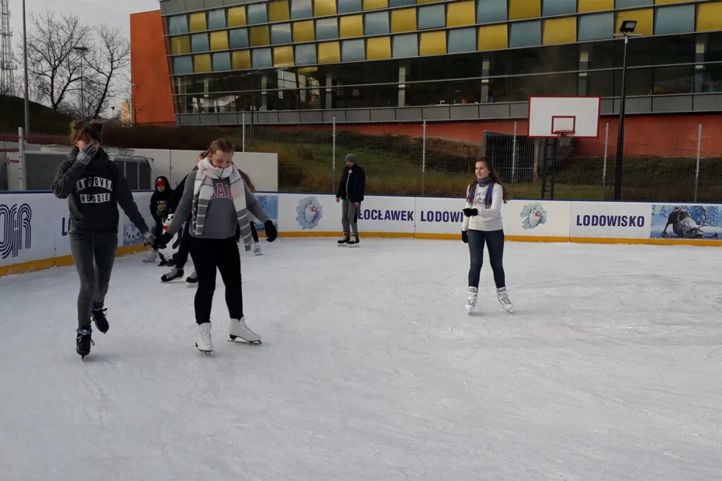 Skaters on the boarded open-air ice of the Wysoka rink on an overcast winter day, with boards reading Włocławek Lodowisko, a basketball hoop and a colourful multi-storey building behind. — Lodowisko Miejskie Wysoka Włocławek