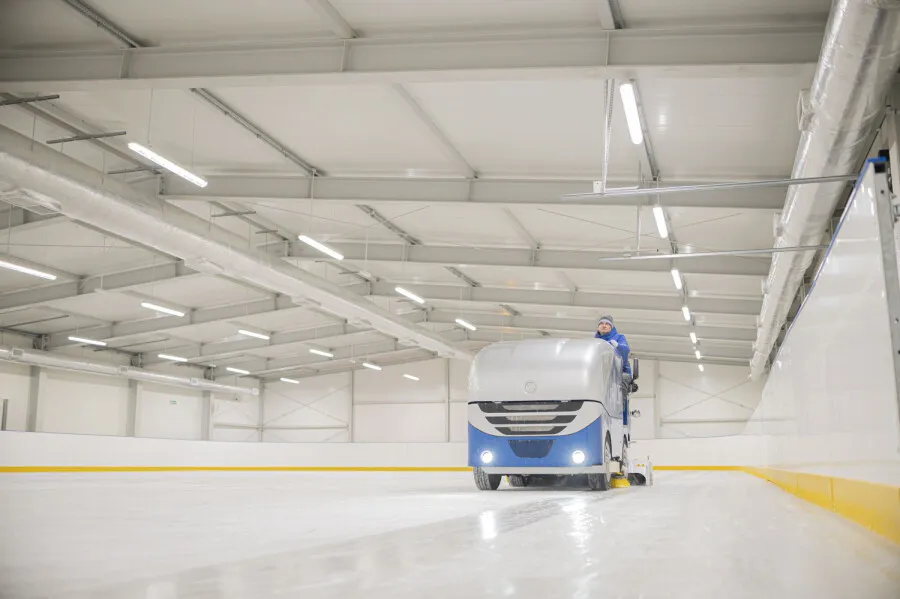 Zamboni jadący wzdłuż bandy pod stalowymi dźwigarami dachu zadaszonego lodowiska w Ełku; po prawej białe bandy z żółtym cokołem. — Lodowisko MOSiR Ełk
