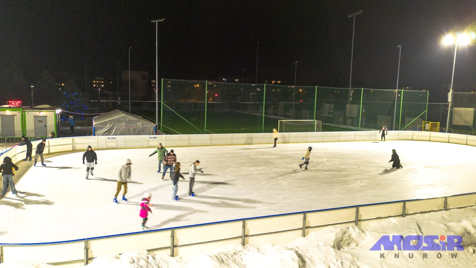 Evening skating on the lit Knurów rink, skaters at a distance behind the boards — Lodowisko Sezonowe MOSiR Knurów