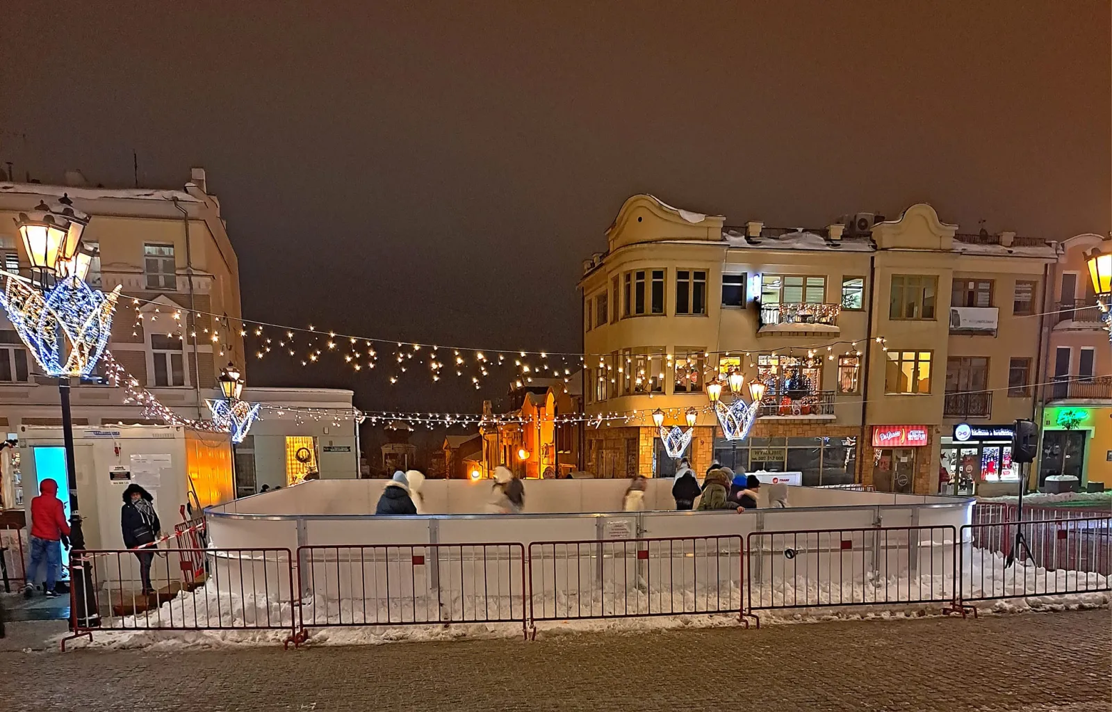 The small boarded rink on the town square in Chełm at night, framed by lit historic townhouses, festive lamp-post decorations and string lights, with snow banked around the barriers. — Lodowisko MOSiR pl. Łuczkowskiego Chełm