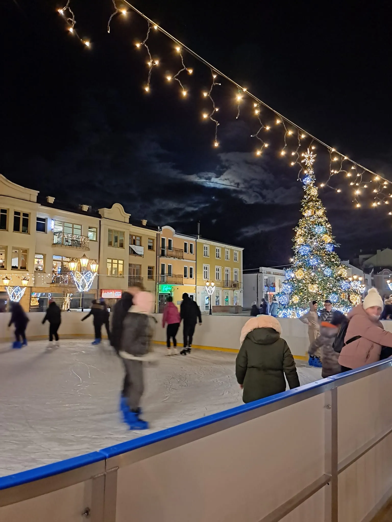 Skaters moving past the white boards of the square rink in Chełm at night, with townhouses and a lit Christmas tree behind. — Lodowisko MOSiR pl. Łuczkowskiego Chełm
