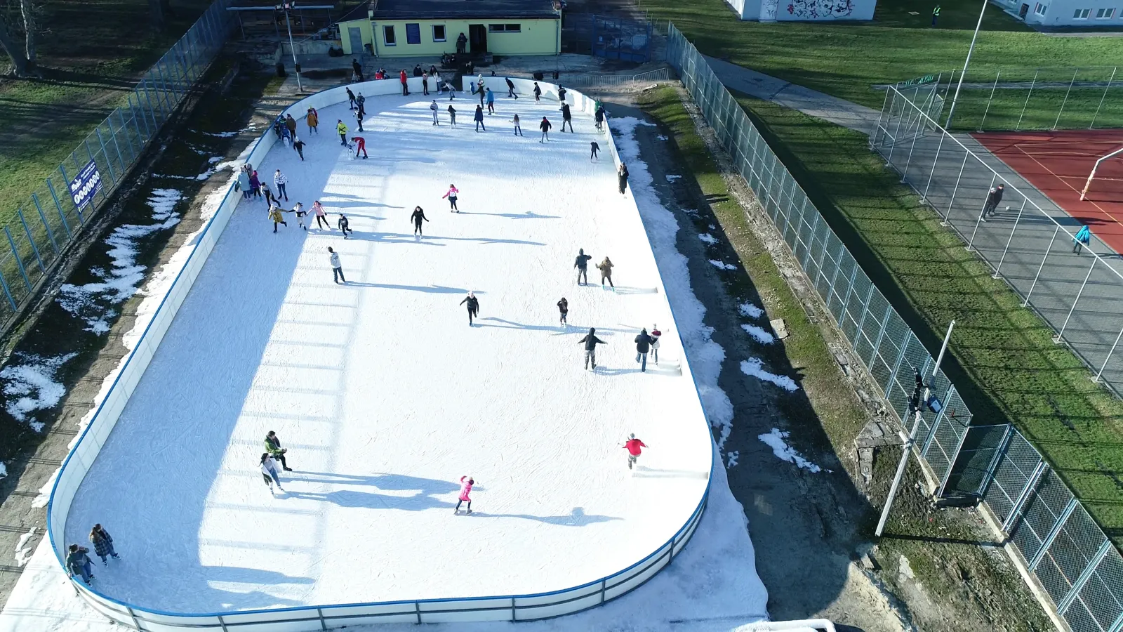 Aerial daytime view of the open-air Sienkiewicza rink in Chełm: a white boarded ice sheet with skaters spread across it, a low yellow building behind, grass and a fenced sports court beside it. — Lodowisko MOSiR ul. Sienkiewicza Chełm