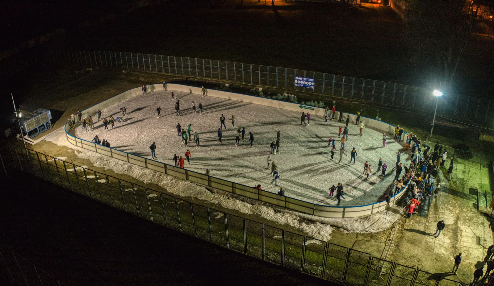Evening aerial view of the floodlit Sienkiewicza ice sheet in Chełm, with many skaters on the ice inside curved white boards and spectators gathered at the edge. — Lodowisko MOSiR ul. Sienkiewicza Chełm