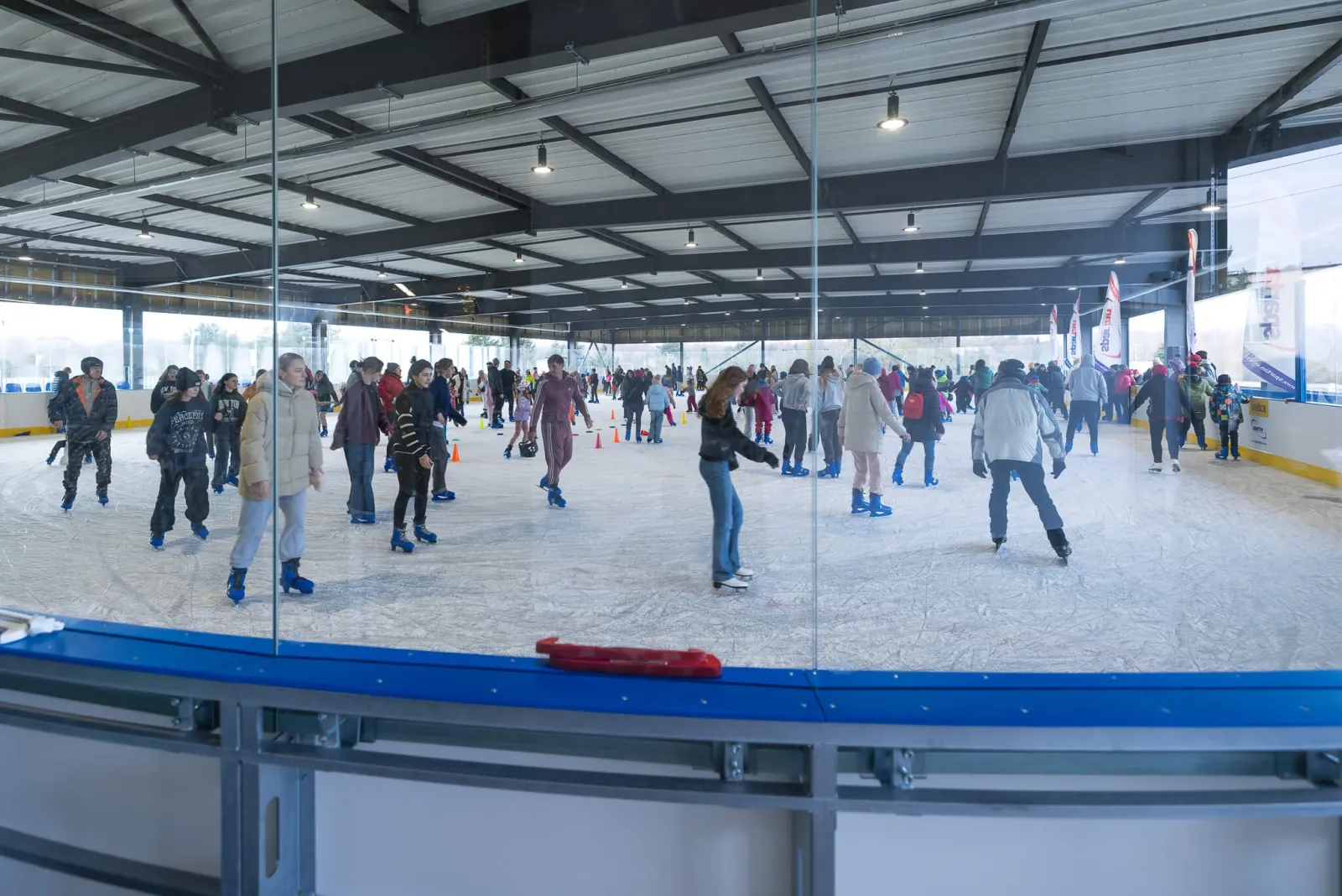 Wide view across the Nowy Dwór ice under the open-sided steel roof, with skaters spread across the rink behind a glass screen. — Lodowisko Nowy Dwór (WCT Spartan)