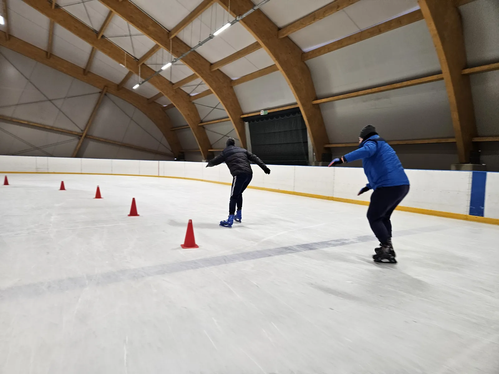 Two skaters weaving past a line of red cones on the boarded ice under the curved laminated-timber roof arches of the Gorlice rink hall. — Lodowisko OSiR Gorlice