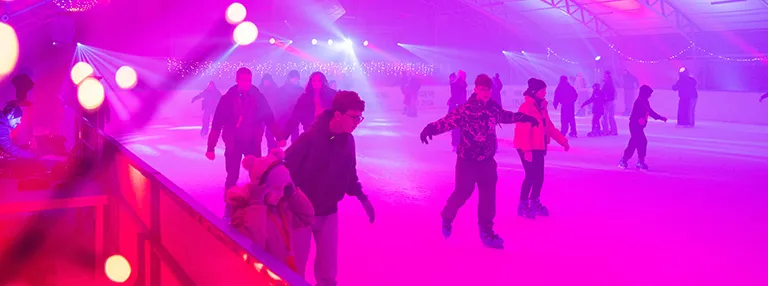 Skaters on the boarded ice of the Suwałki municipal rink during a disco session, lit in pink and magenta with spotlights and strings of lights under the roof. — Lodowisko OSiR Suwałki (przy SP nr 11)