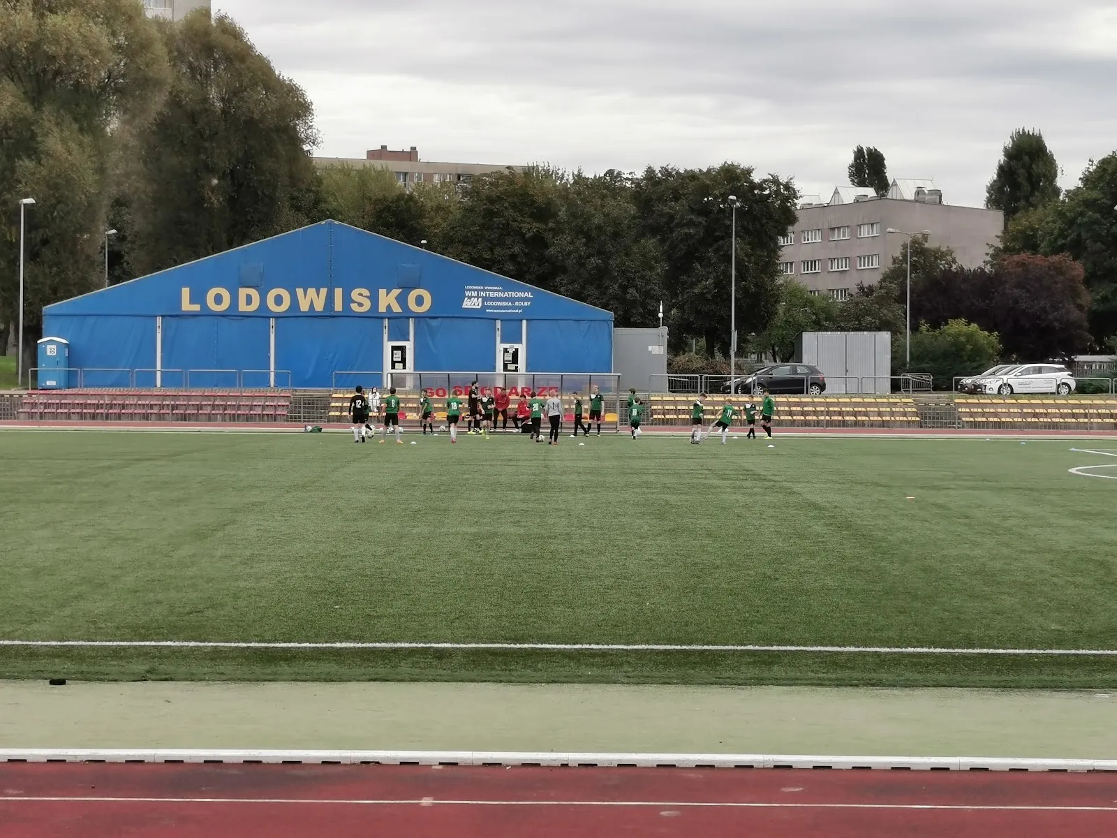 Niebieski namiot z napisem „Lodowisko” obok stadionu lekkoatletycznego, widziany zza boiska piłkarskiego. — Lodowisko OSiR Targówek (Łabiszyńska)