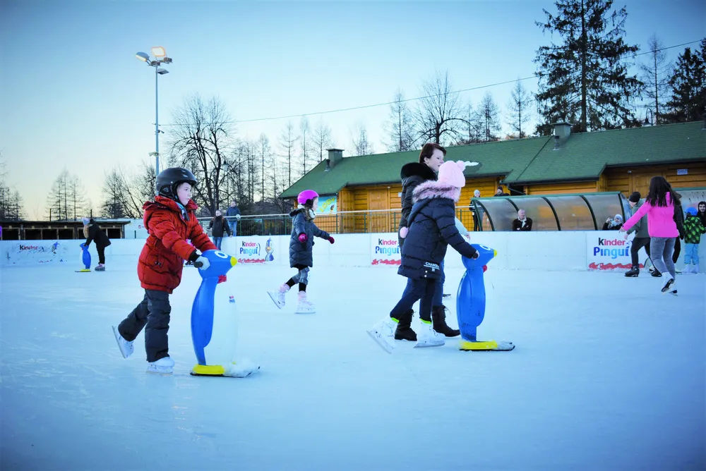The Brenna ice rink beside the timber park building, with children and adults skating in the winter light. — Lodowisko w Parku Turystyki (Brenna)