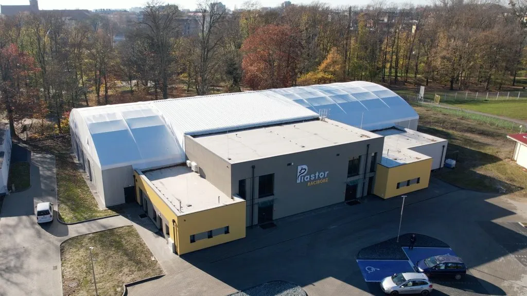 Widok z drona na budynek lodowiska Piastor w Raciborzu: długa biała hala z dachem namiotowym obok szaro-żółtego bloku wejściowego z napisem Piastor, wśród jesiennych drzew i parkingu. — Lodowisko Piastor Racibórz
