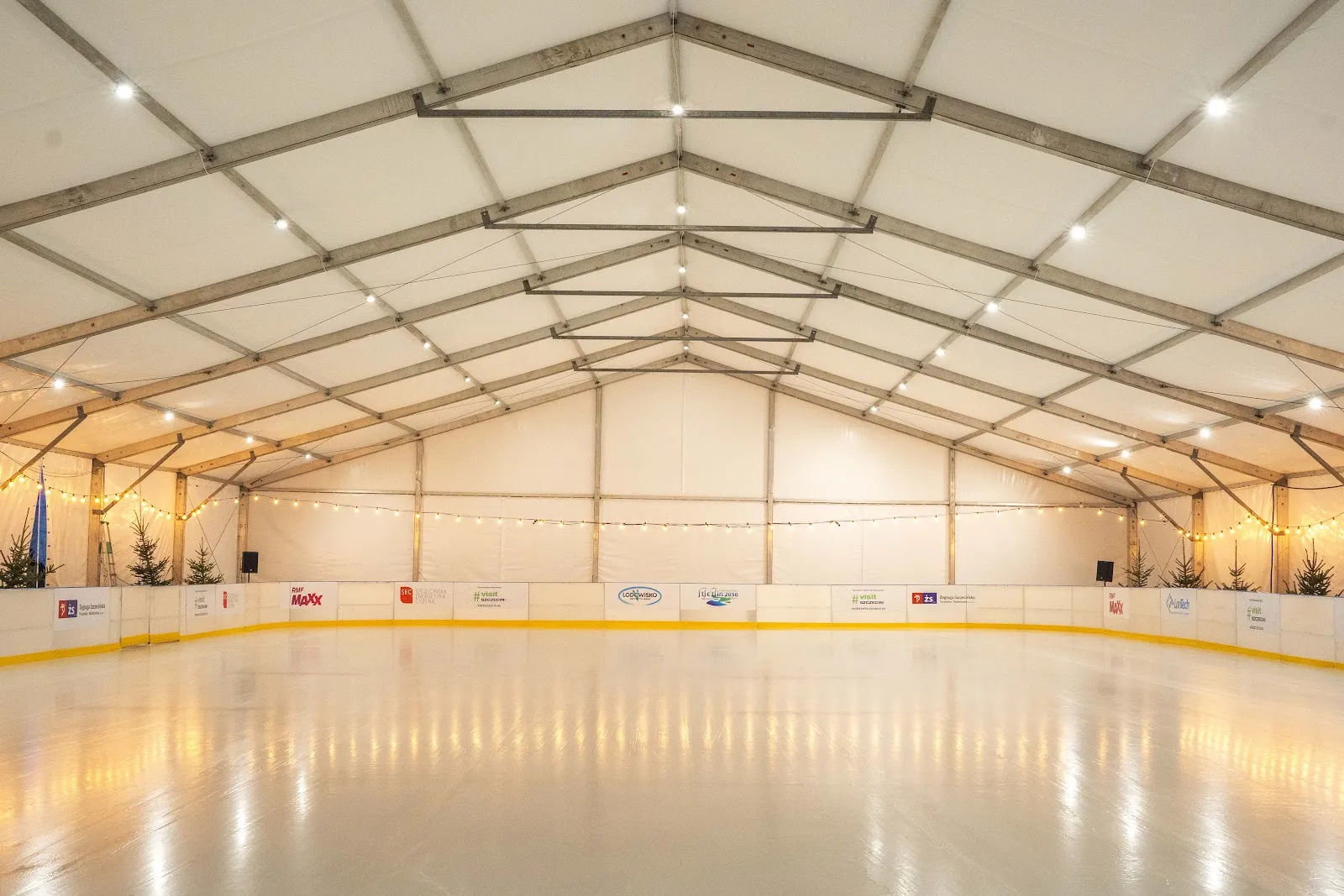 Empty ice under the white tent hall with fairy lights along the walls and boards carrying sponsor panels. — Lodowisko przy Arenie