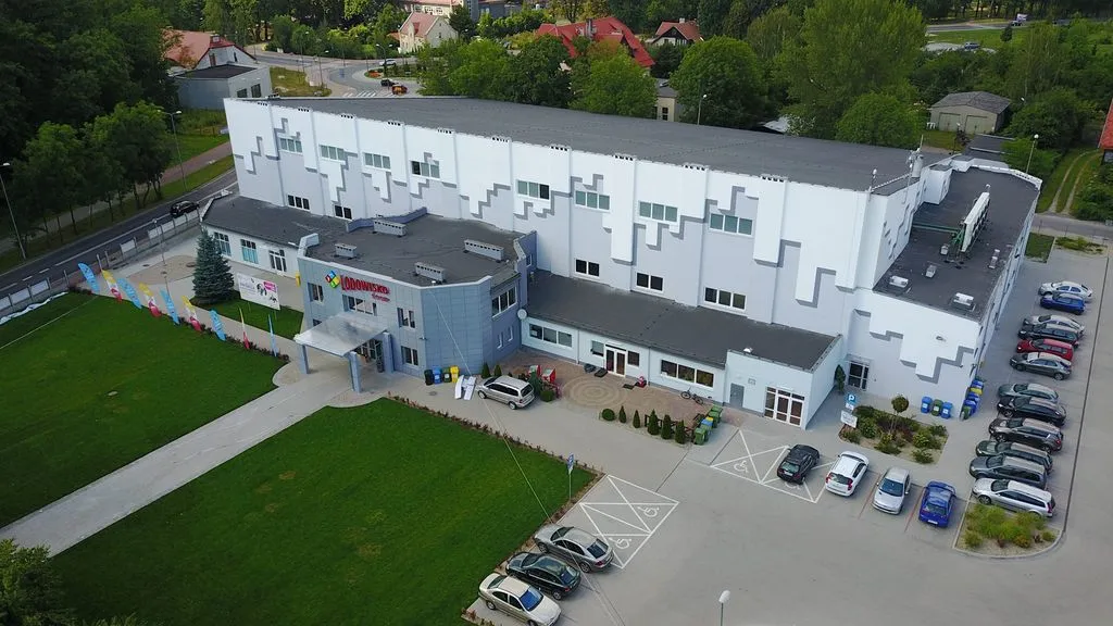 Aerial view of the Świdnica ice hall: a large white and grey building with the Lodowisko sign over a glass entrance, a paved forecourt with disabled parking bays, a lawn with banners and a car park beside it. — Lodowisko ŚOSiR Świdnica