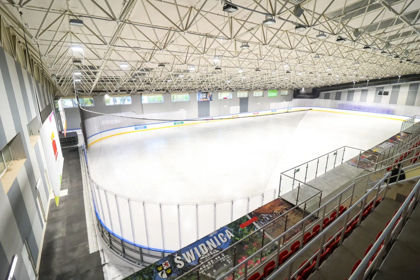 The 60 x 30 m ice sheet seen from the stands in the Świdnica hall, with red seats along the right, boards with a Świdnica banner and a steel-truss roof overhead. — Lodowisko ŚOSiR Świdnica