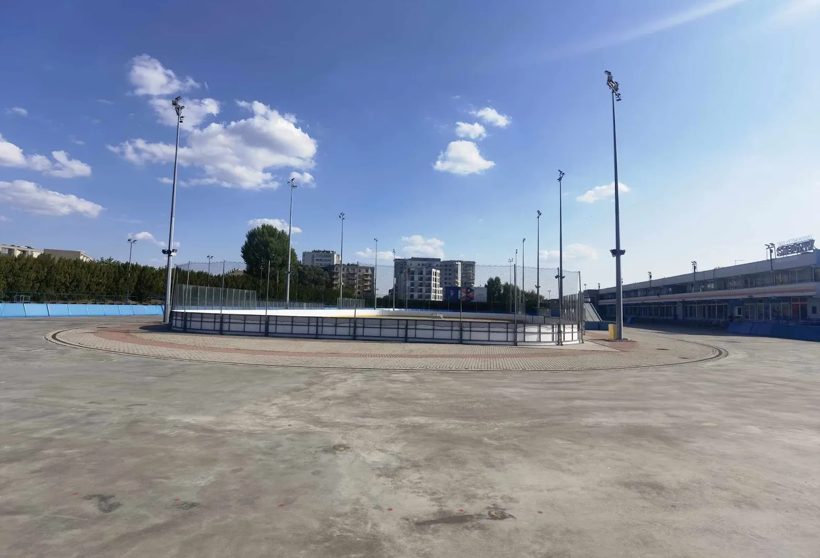 Pusty tor na Stegnach latem, z bandami lodowiska hokejowego, masztami reflektorów i budynkiem trybun po prawej. — Lodowisko Stegny (Aktywna Warszawa)