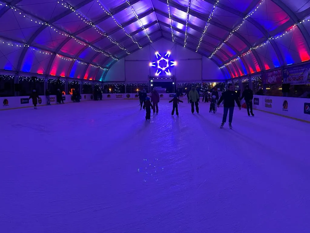 Skaters on the covered, colour-lit ice of the Szwajcaria Bałtowska rink under the arched tent roof — Lodowisko Szwajcaria Bałtowska