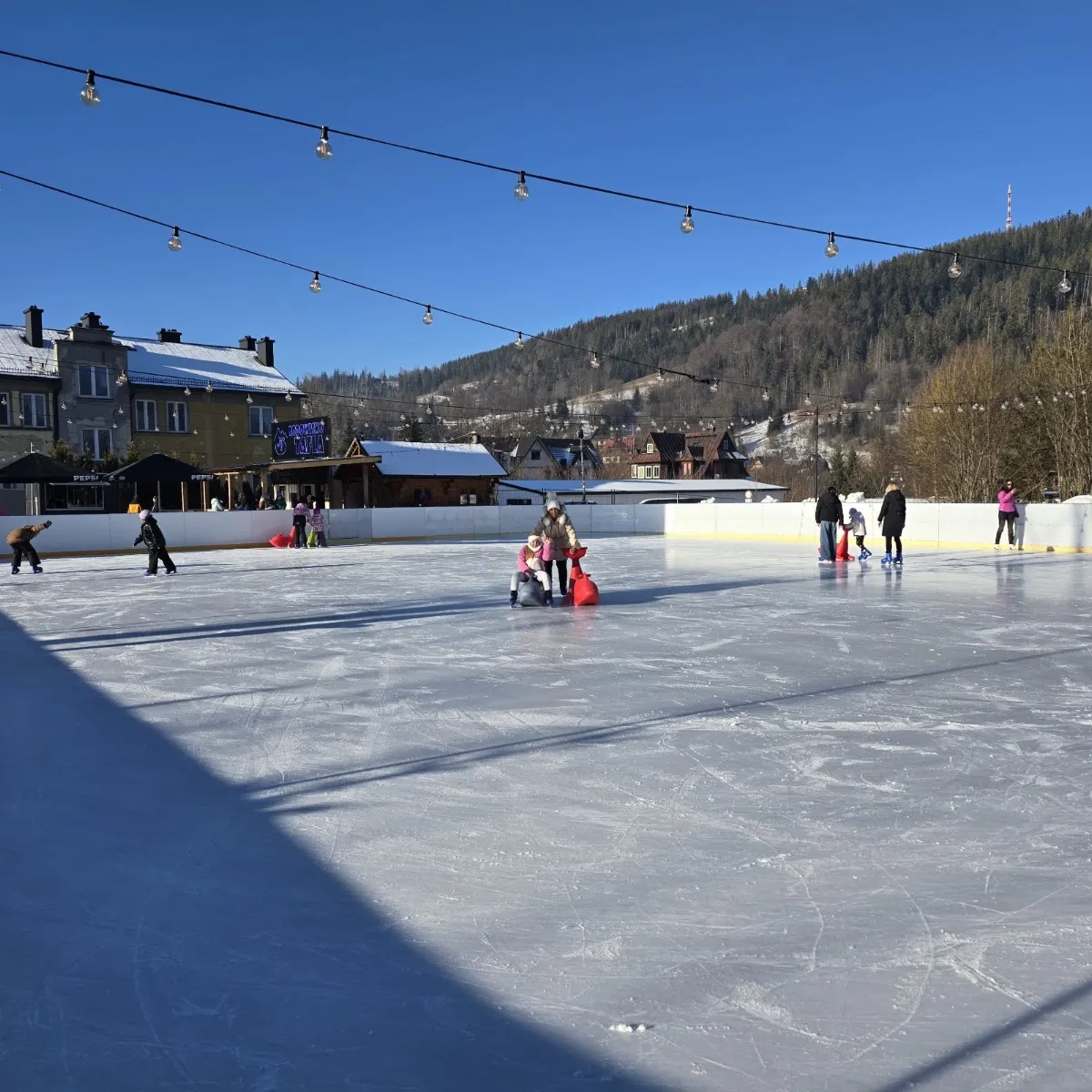 The open-air ice of Lodowisko Tafla in Zakopane on a sunny day, skaters at a distance with the Tatra slopes behind — Lodowisko TAFLA (Zakopane)